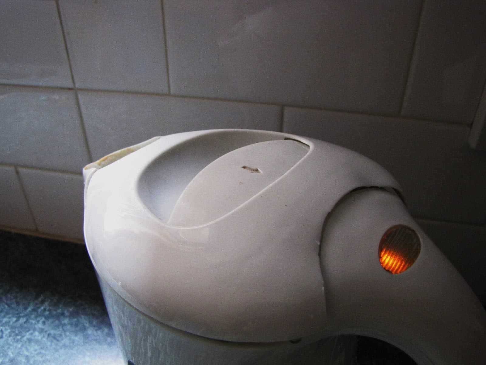 A white cordless electric kettle with its power light on, against kitchen tiles